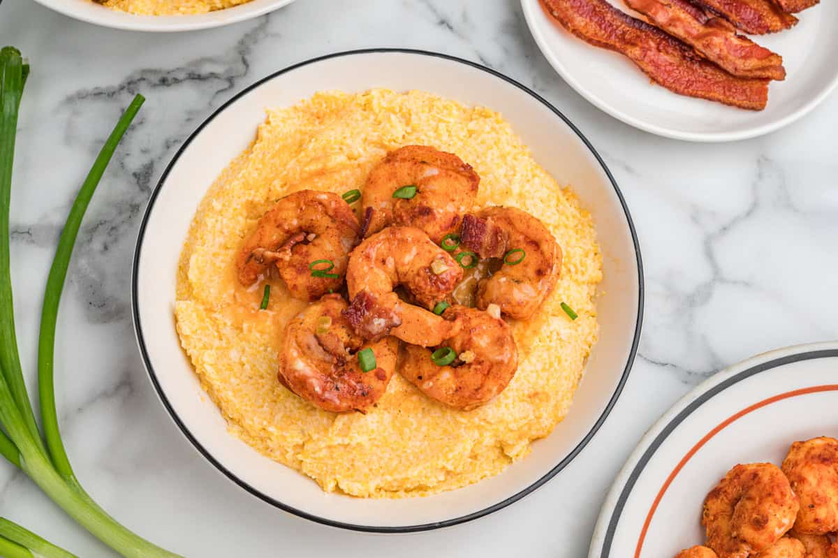 Bowl of cheesy grits topped with shrimp, bacon pieces, and scallions. The bowl is surrounded by fresh green onions and a plate of bacon.