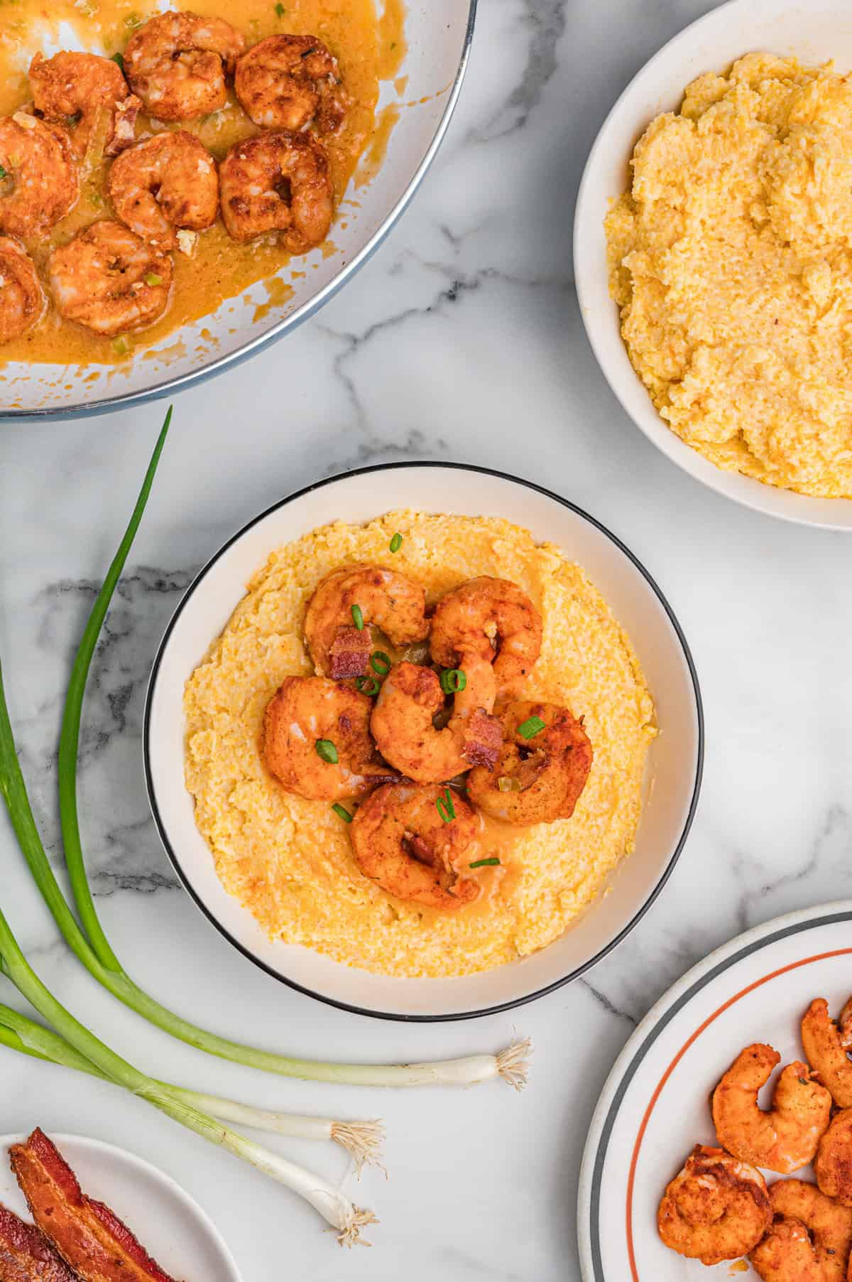 Overhead view of a bowl of cheesy shrimp and grits with bacon and green onions. Surrounding it are a skillet with saucy shrimp, a bowl of cheesy grits, a plate of cooked shrimp, fresh green onions, and a plate of bacon, all arranged on a marble surface.
