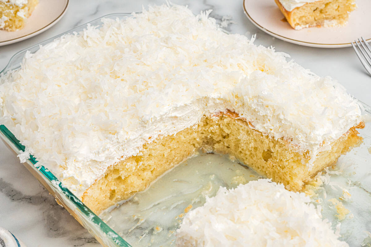 A baked coconut poke cake in a glass dish with several slices missing. The cake has a fluffy white whipped topping and is generously sprinkled with shredded coconut.
