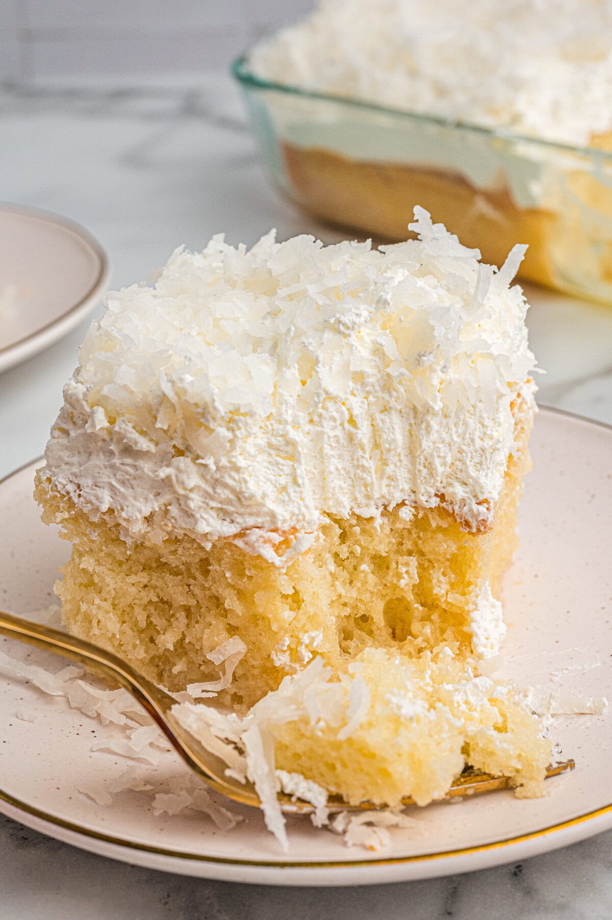 A close-up of a coconut poke cake slice on a plate with a bite taken out and placed on a fork in the foreground. The cake shows a moist, spongy texture and a thick whipped topping with shredded coconut.