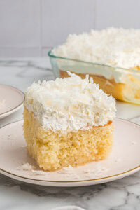 A tall slice of coconut poke cake on a plate, showing its fluffy base, thick whipped topping, and coconut sprinkled on top.