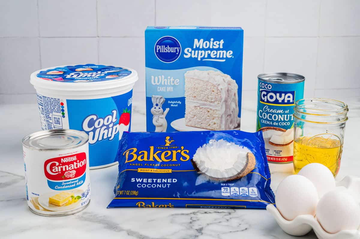 Ingredients for a coconut poke cake, including a tub of Cool Whip, a box of Pillsbury Moist Supreme White Cake Mix, a can of Goya cream of coconut, a can of Carnation sweetened condensed milk, a bag of Baker's sweetened coconut flakes, three eggs, and oil.