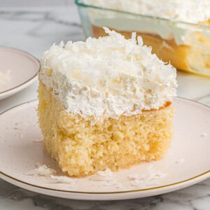 A square slice of coconut poke cake on a white plate with a fluffy whipped topping and shredded coconut. The full cake in a baking dish is visible in the background.
