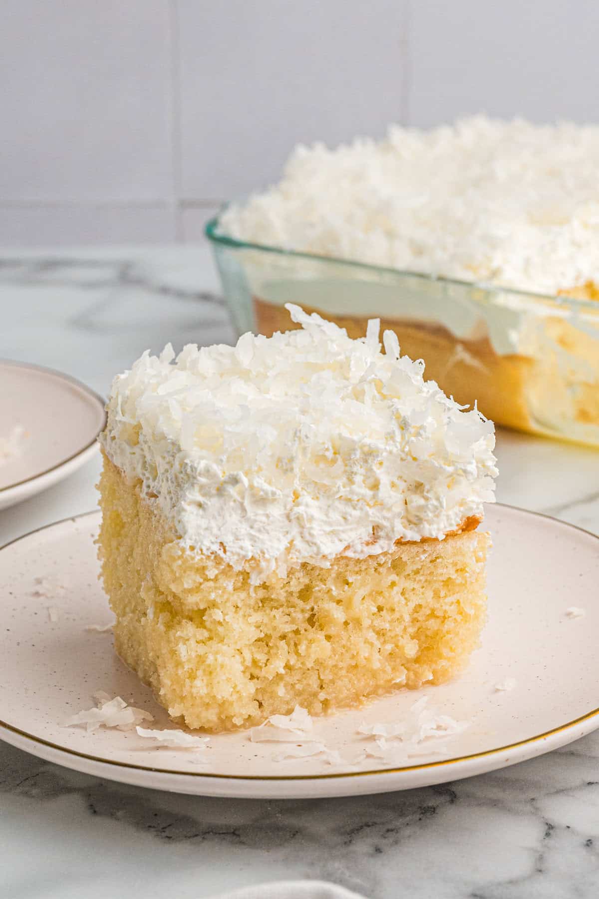A tall slice of coconut poke cake on a plate, showing its fluffy base, thick whipped topping, and coconut sprinkled on top.