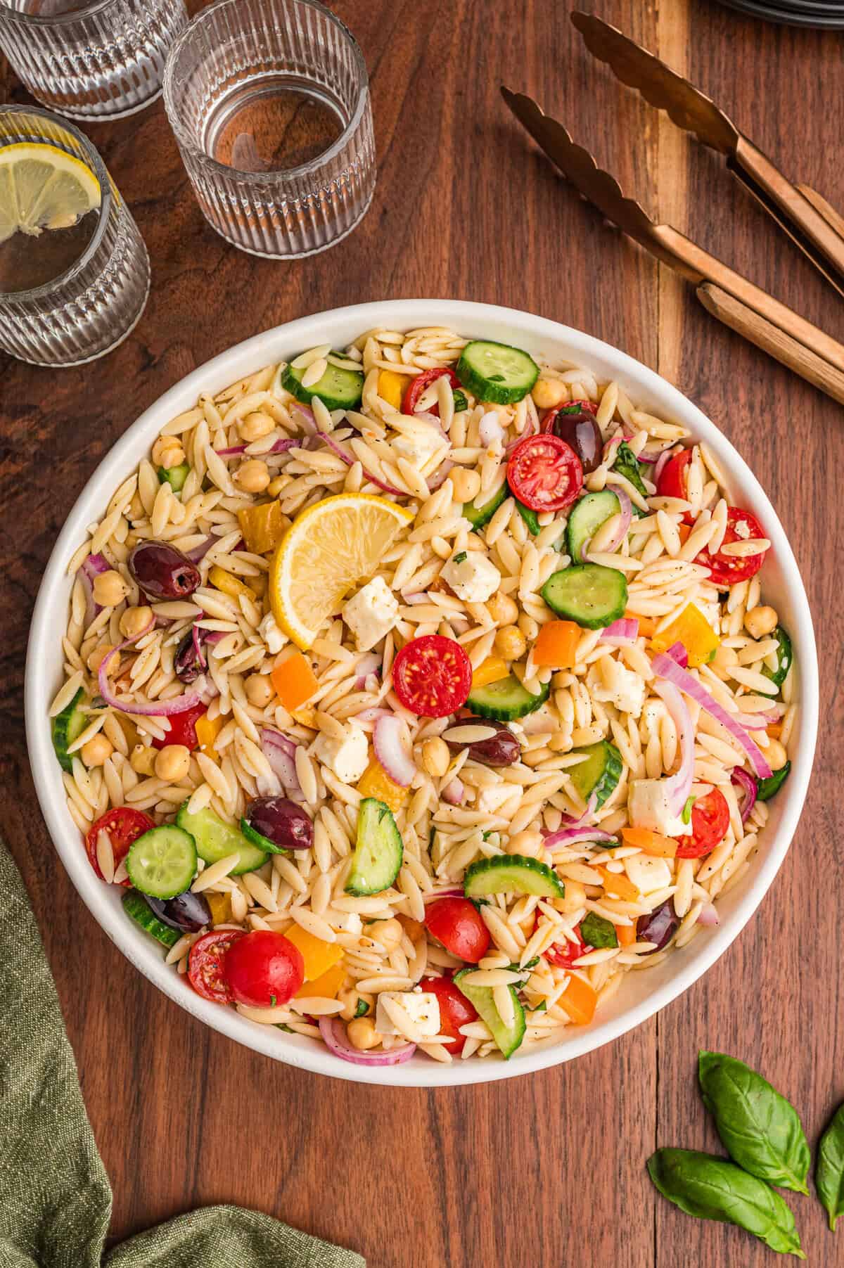 Overhead view of a large white bowl filled with orzo salad containing sliced cucumbers, cherry tomatoes, bell peppers, red onions, chickpeas, kalamata olives, feta cheese, and lemon garnish, placed on a wooden table with clear glasses of lemon water and bronze utensils in the background.
