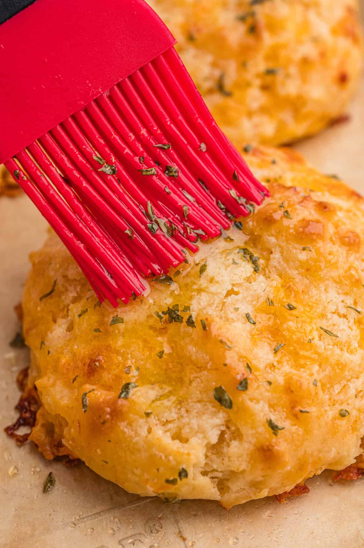 A red silicone brush applies melted butter with herbs onto the top of a freshly baked cheddar biscuit, enhancing its golden, textured surface.