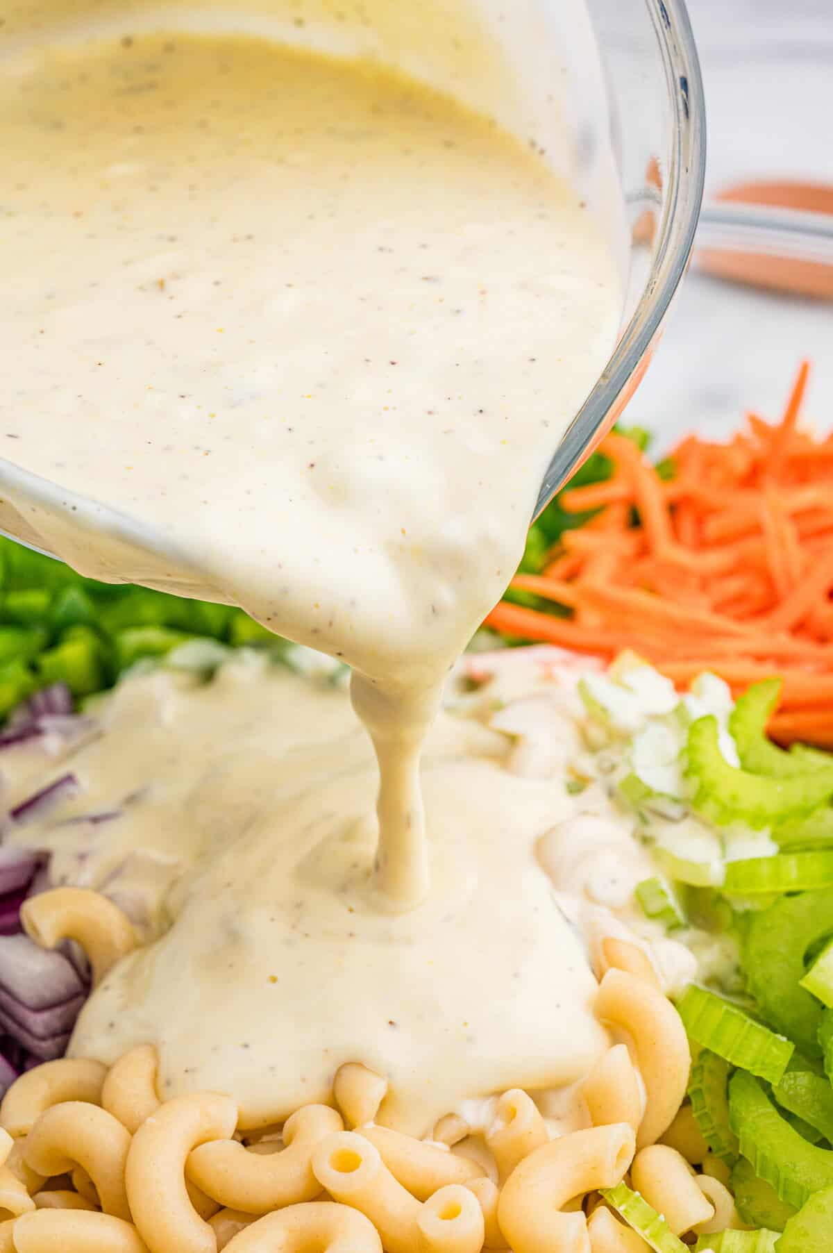 Creamy dressing is being poured from a glass bowl over a mixture of uncooked elbow macaroni, chopped red onion, green bell pepper, shredded carrots, and celery.