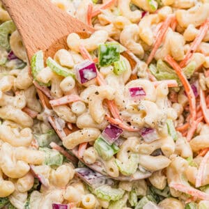 Close-up of a wooden spoon lifting a serving of old fashioned macaroni salad, showing elbow macaroni, shredded carrots, diced celery, green bell pepper, and red onion in a creamy dressing.