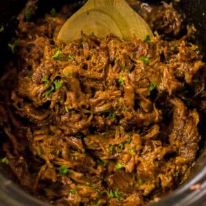 Close-up of shredded BBQ beef in a slow cooker, mixed with sauce and garnished with chopped green herbs. A wooden spoon is partially visible at the top.