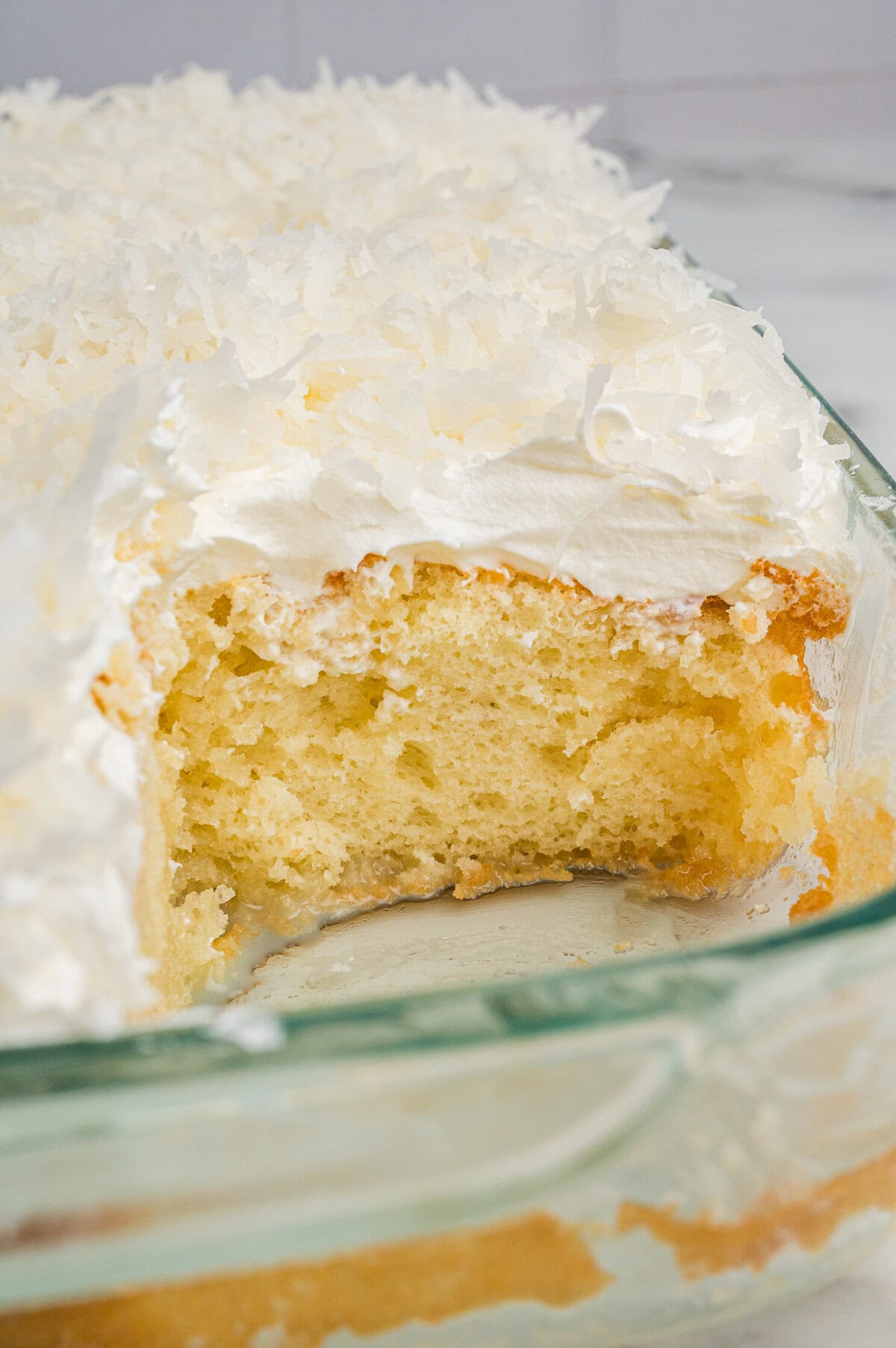 A close-up view of a moist yellow coconut poke cake with a square slice removed, revealing its soft texture. The cake is topped with fluffy whipped topping and shredded coconut in a glass baking dish.