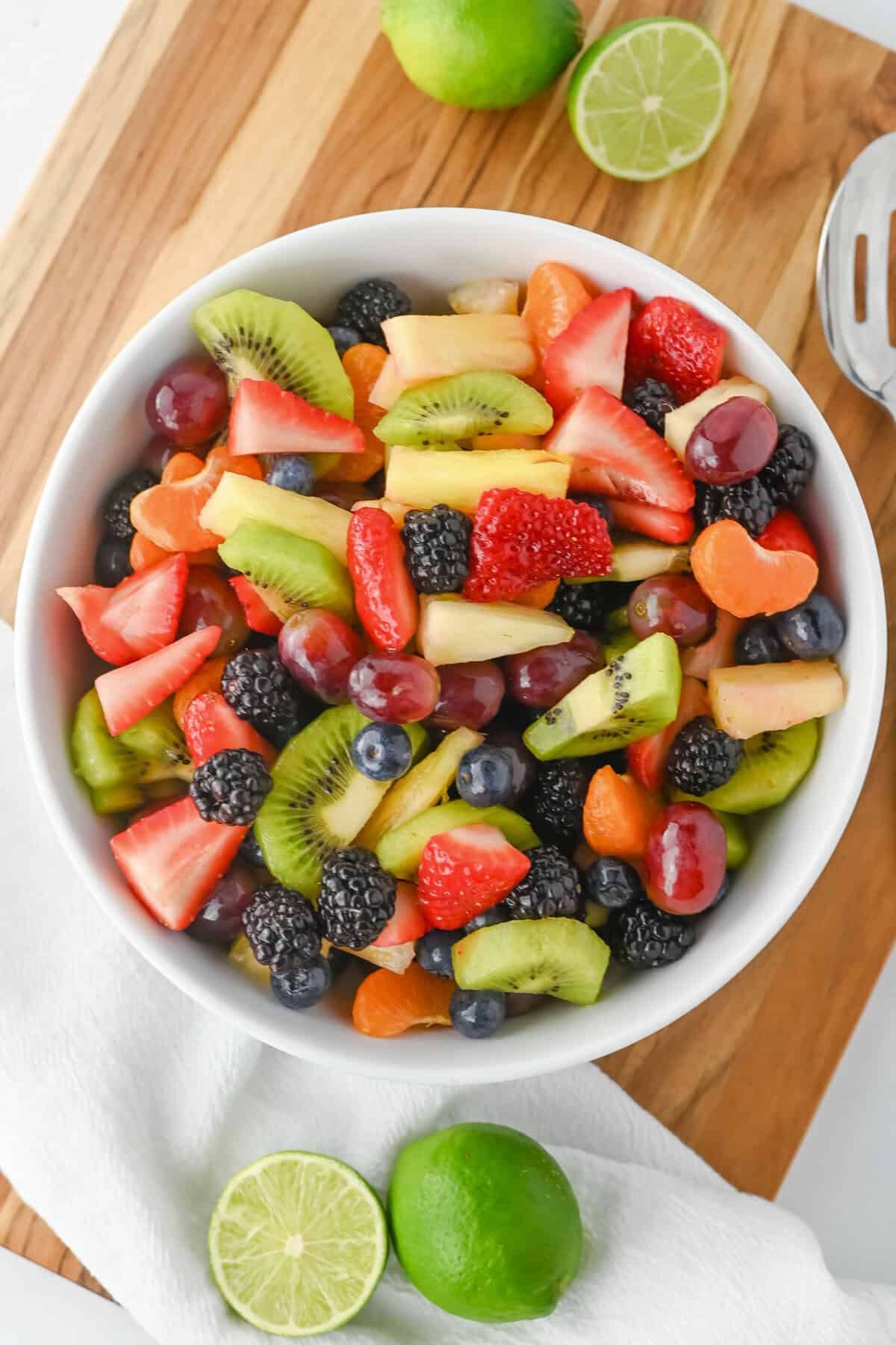Fruit salad with honey lime dressing in a white bowl with sliced strawberries, kiwi, pineapple, grapes, blackberries, blueberries, and clementine slices. The bowl is set on a wooden board with halved limes nearby.