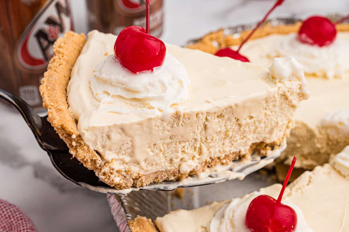 A slice of root beer float pie being lifted from the pan, showing creamy filling, graham cracker crust, and a topping of whipped cream and a cherry.