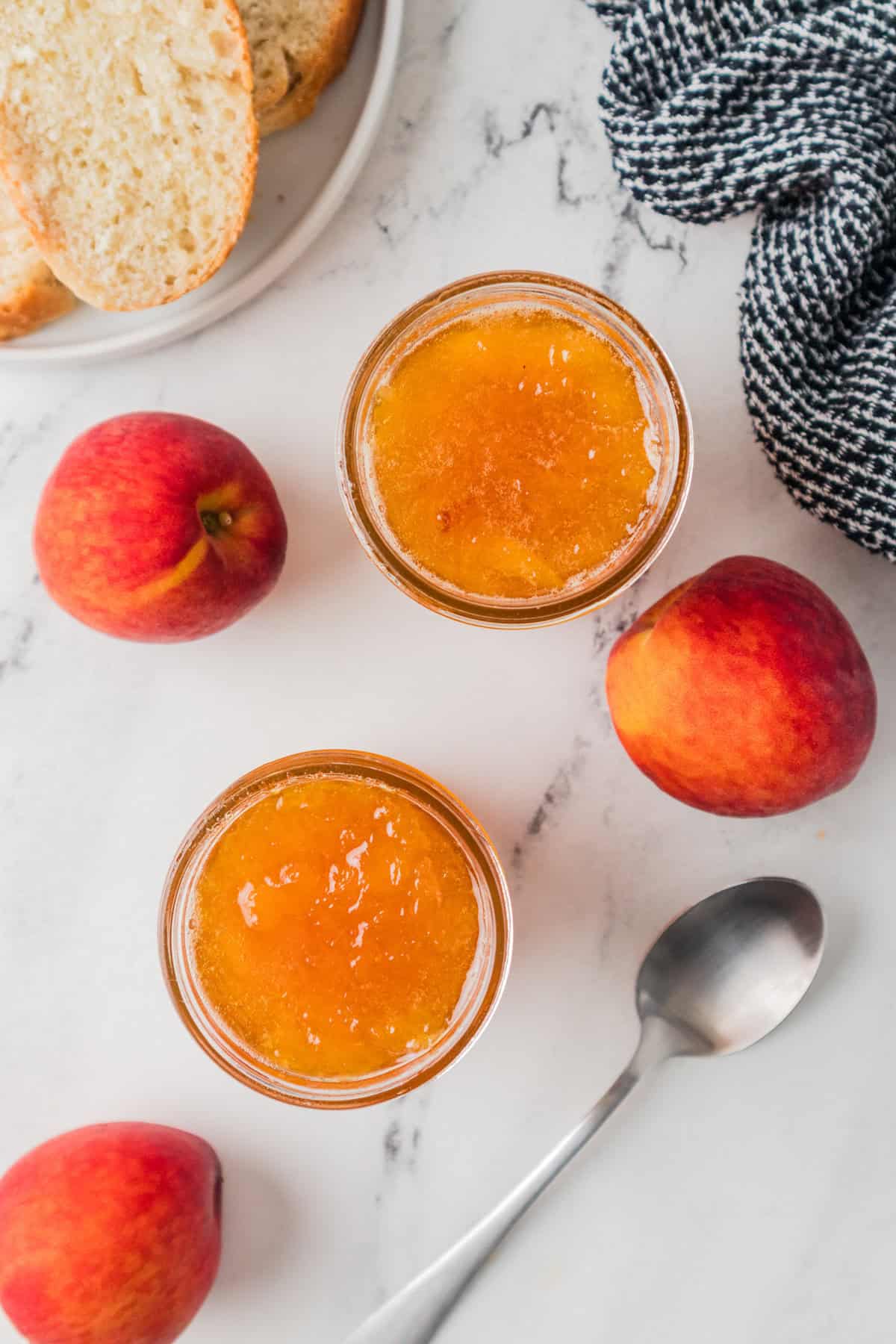 Overhead view showing two open jars of peach jam on a marble surface, surrounded by whole fresh peaches, a silver spoon, a black-and-white cloth, and a plate with slices of bread.