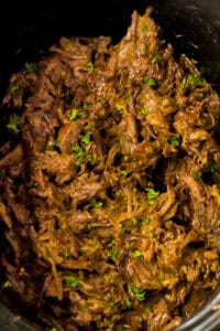 Overhead image showing slow cooker BBQ beef filling the slow cooker, mixed with BBQ sauce and sprinkled with fresh chopped herbs.