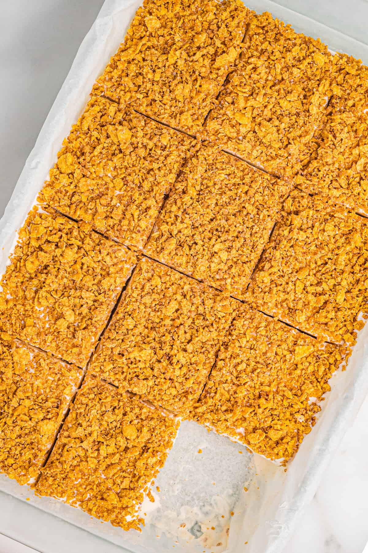 Overhead view of a parchment-lined pan filled with fried ice cream dessert cut into even square portions. Each square has a crunchy golden cornflake topping, and one bar has been removed, revealing the creamy interior and base layer.