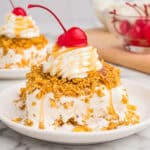 Fried ice cream cake on a white plate, coated in a crispy golden crust, topped with whipped cream, caramel drizzle, and a maraschino cherry.