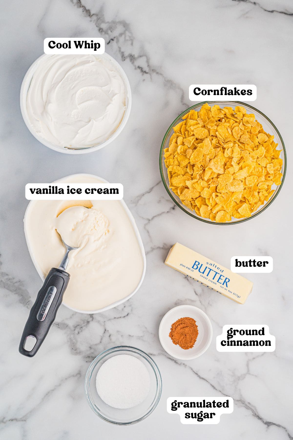 Ingredients for fried ice cream arranged on a marble surface, each labeled. Includes a tub of Cool Whip, a bowl of cornflakes, a container of vanilla ice cream with a scoop, a stick of butter, a small dish of ground cinnamon, and a small bowl of granulated sugar.