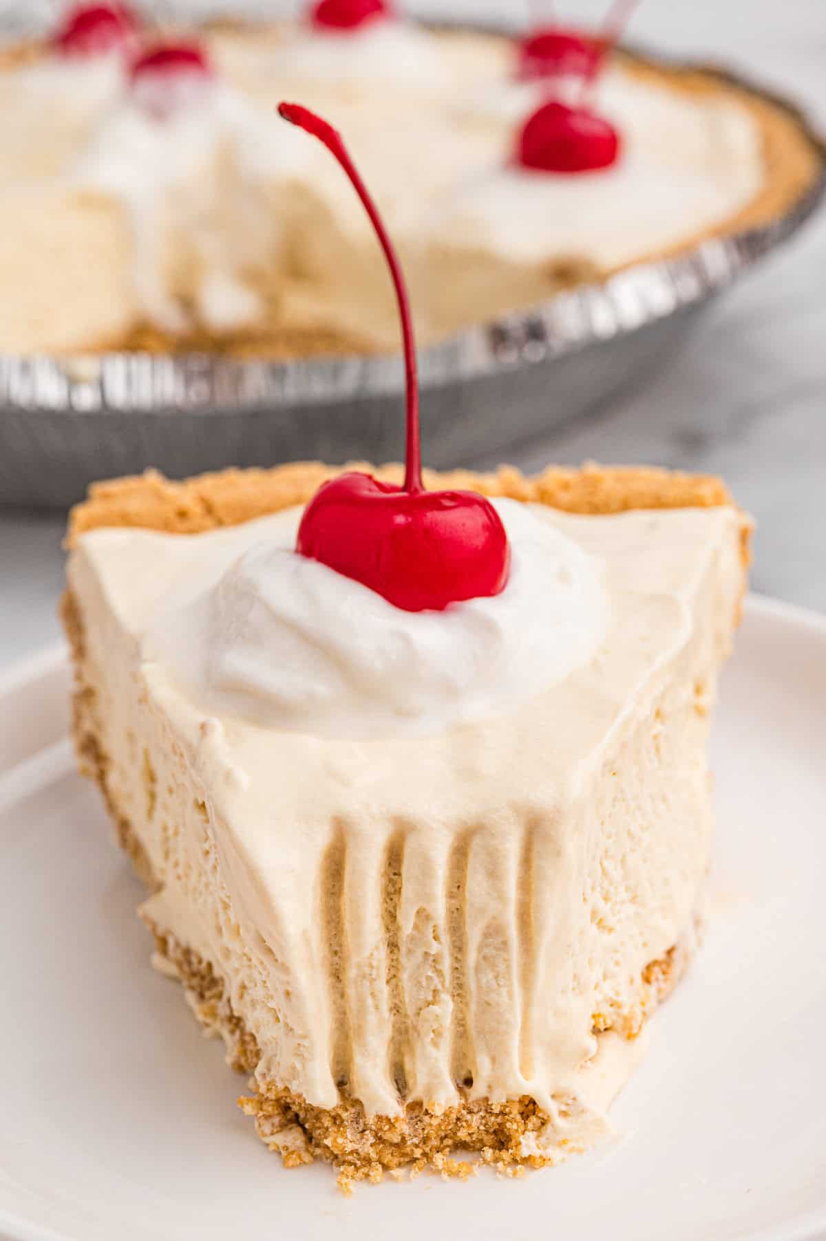 A close-up of a plated slice of root beer float pie with a graham cracker crust. The pie slice has creamy, slightly melted filling with visible fork marks and is topped with whipped cream and a maraschino cherry.