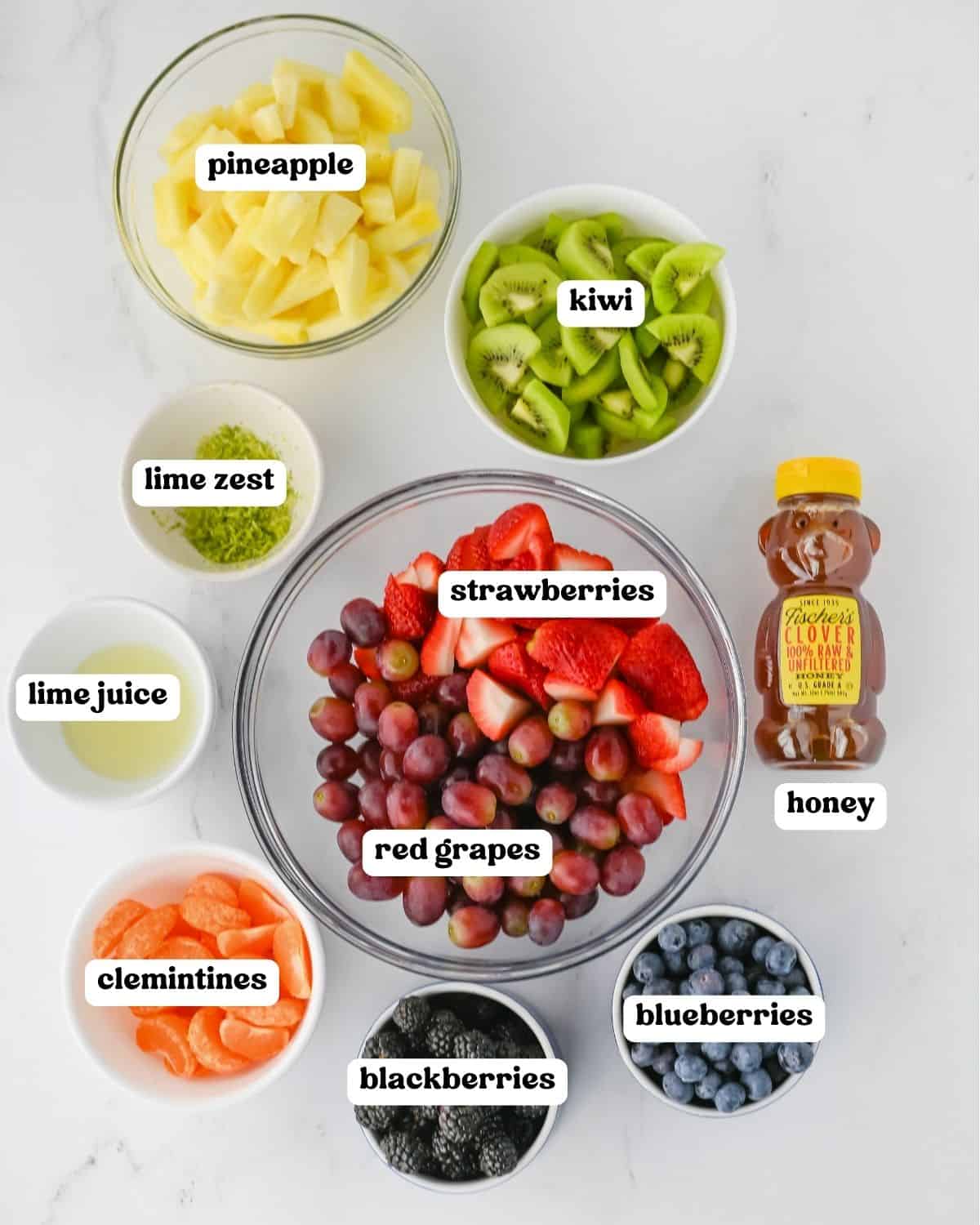 Fruit salad ingredients including pineapple, kiwi, strawberries, red grapes, blueberries, blackberries, clementines, lime zest, lime juice, and honey.
