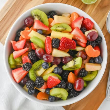 Fruit salad with honey lime dressing in a white bowl with sliced strawberries, kiwi, pineapple, grapes, blackberries, blueberries, and clementine slices.