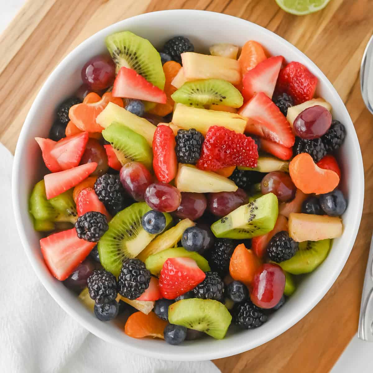 Fruit salad with honey lime dressing in a white bowl with sliced strawberries, kiwi, pineapple, grapes, blackberries, blueberries, and clementine slices.