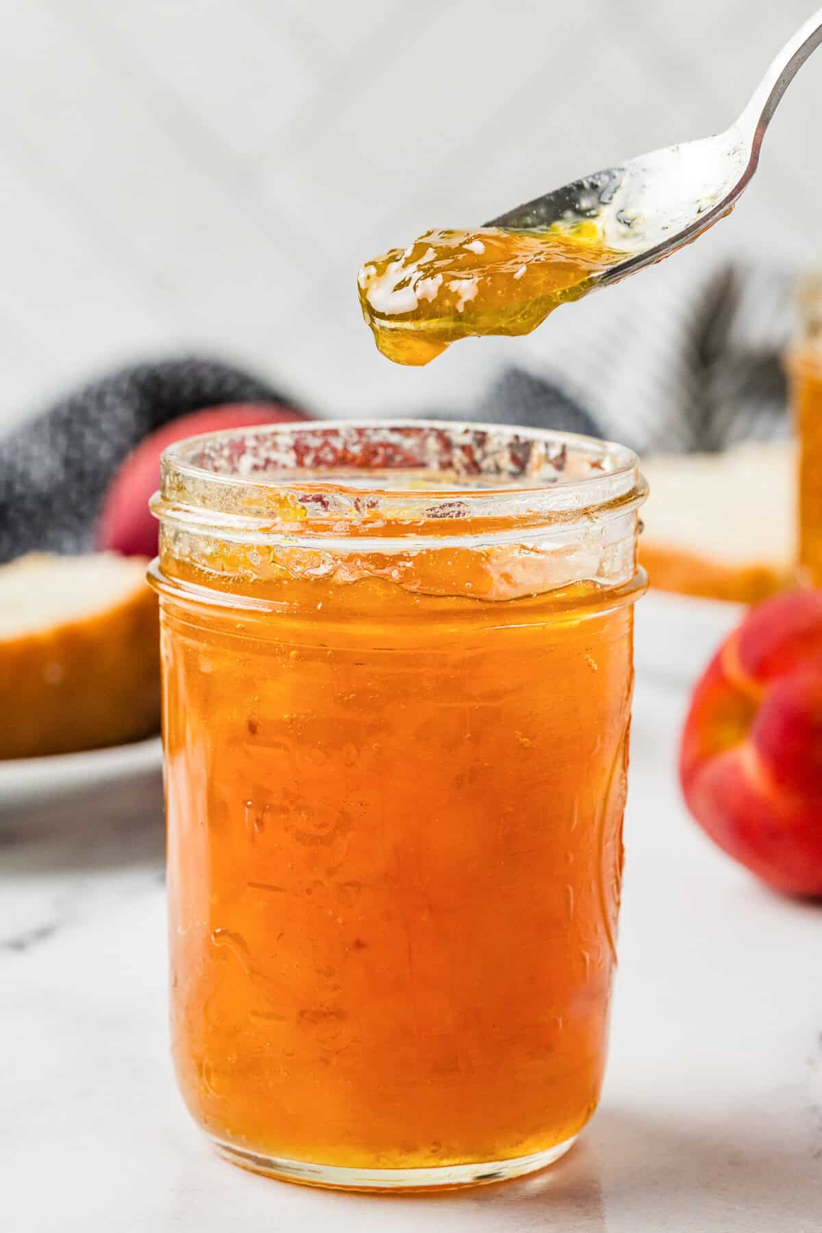 Close-up of a spoon lifting thick peach jam from an open mason jar, with a peach and bread in the background.