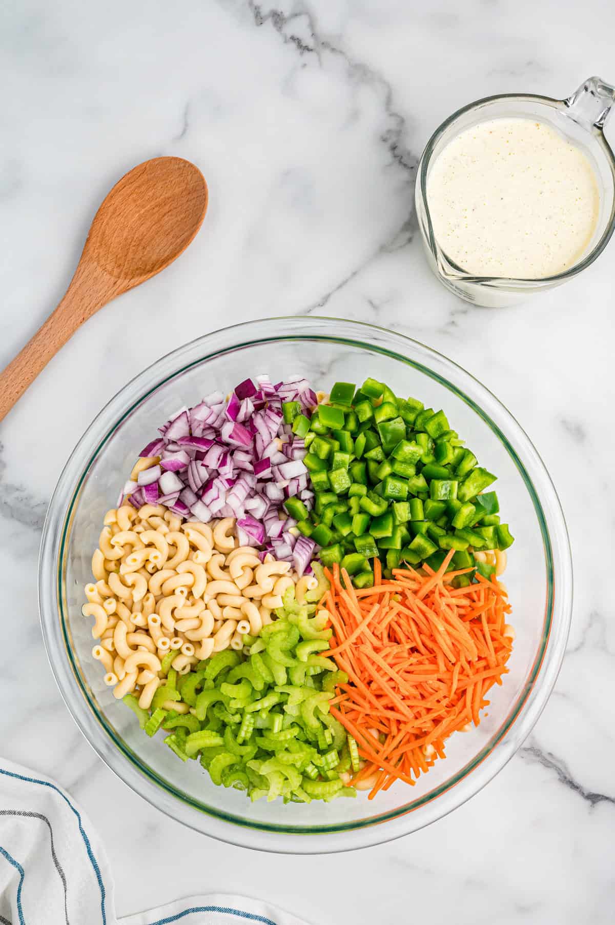 Overhead view of a glass bowl filled with separated ingredients for macaroni salad: diced red onion, green bell pepper, shredded carrots, sliced celery, and elbow macaroni. A wooden spoon and a measuring cup of creamy dressing sit nearby on a marble countertop.