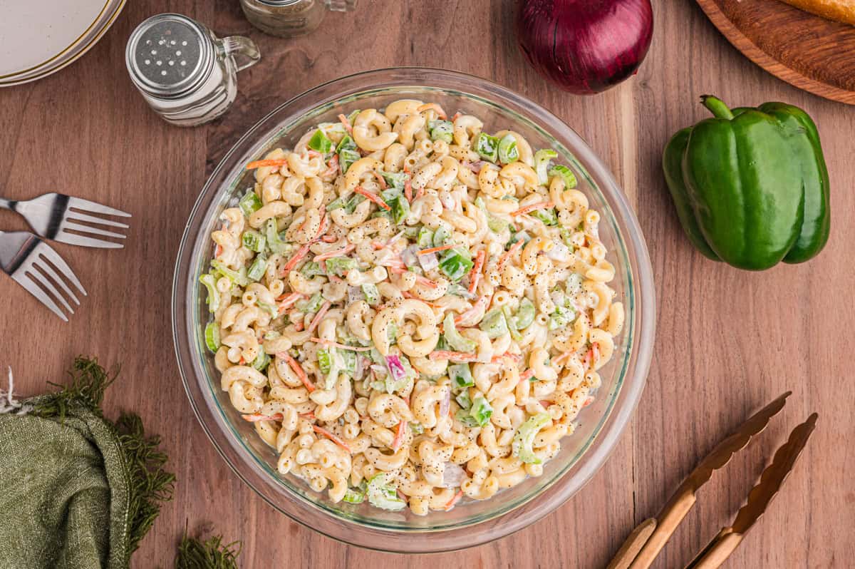 Overhead shot of a glass bowl of macaroni pasta salad placed on a wooden table with surrounding items including forks, seasoning jars, a red onion, a green bell pepper, and tongs.