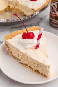 A slice of root beer float pie on a white plate, topped with whipped cream, a cherry, and a red-striped paper straw.