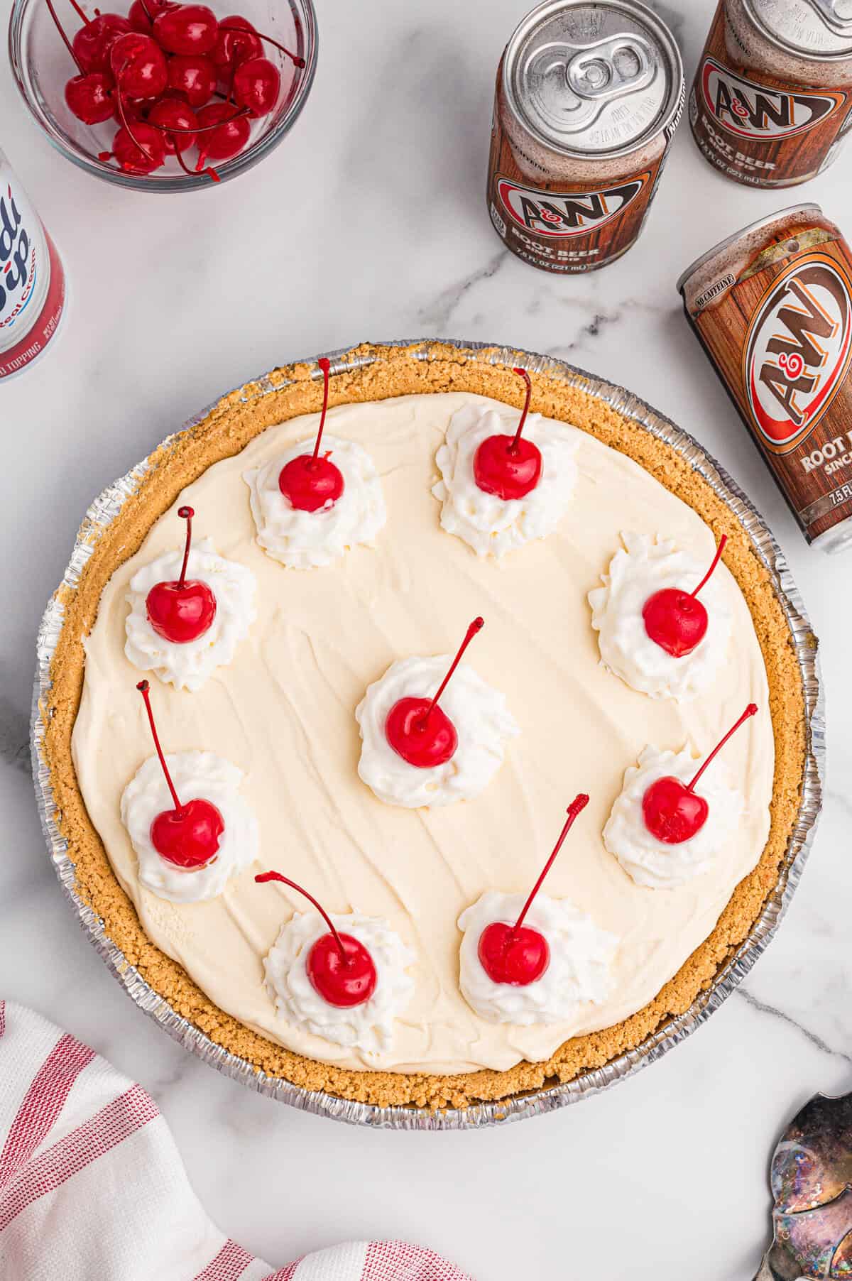 Overhead view of a full root beer float pie with whipped cream dollops and cherries; surrounded by root beer cans and a bowl of cherries.