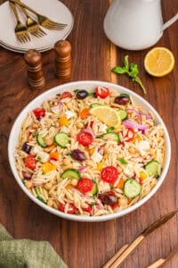 A bowl filled with orzo salad, highlighting visible ingredients such as halved cherry tomatoes, cucumber slices, red onion, chickpeas, yellow and orange bell pepper pieces, kalamata olives, cubed feta, and chopped herbs, with a lemon wedge on top.