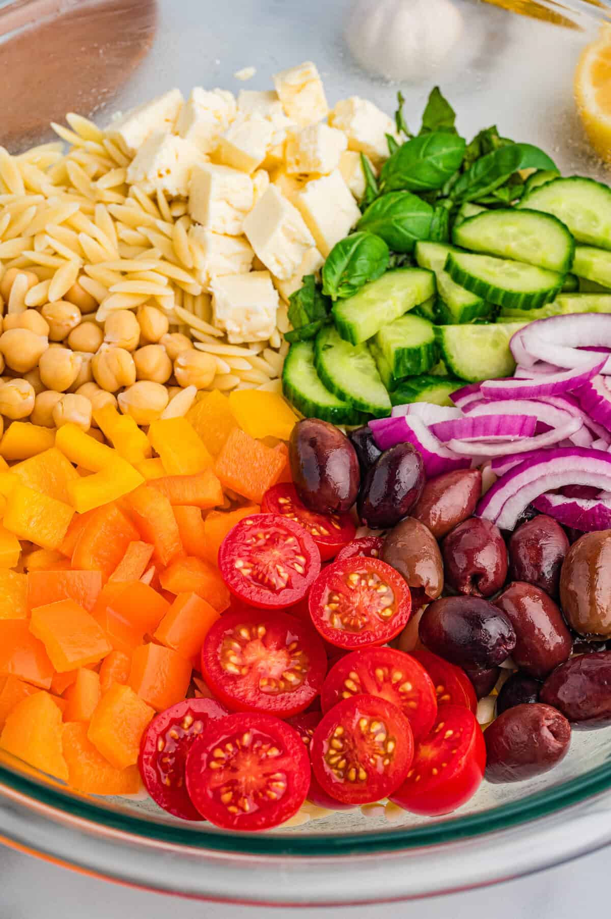 lose-up image of a glass mixing bowl with orzo salad ingredients placed in neat sections, including cherry tomatoes, kalamata olives, orange and yellow bell peppers, chickpeas, feta, basil, cucumber, and red onion.