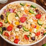 A bowl filled with orzo salad, highlighting visible ingredients such as halved cherry tomatoes, cucumber slices, red onion, chickpeas, yellow and orange bell pepper pieces, kalamata olives, cubed feta, and chopped herbs, with a lemon wedge on top.