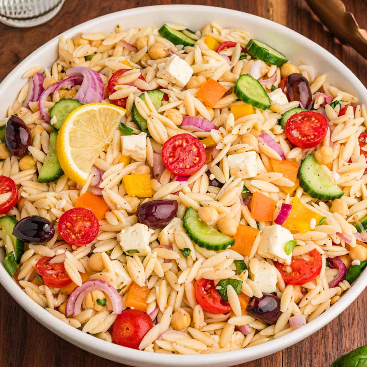 A bowl filled with orzo salad, highlighting visible ingredients such as halved cherry tomatoes, cucumber slices, red onion, chickpeas, yellow and orange bell pepper pieces, kalamata olives, cubed feta, and chopped herbs, with a lemon wedge on top.