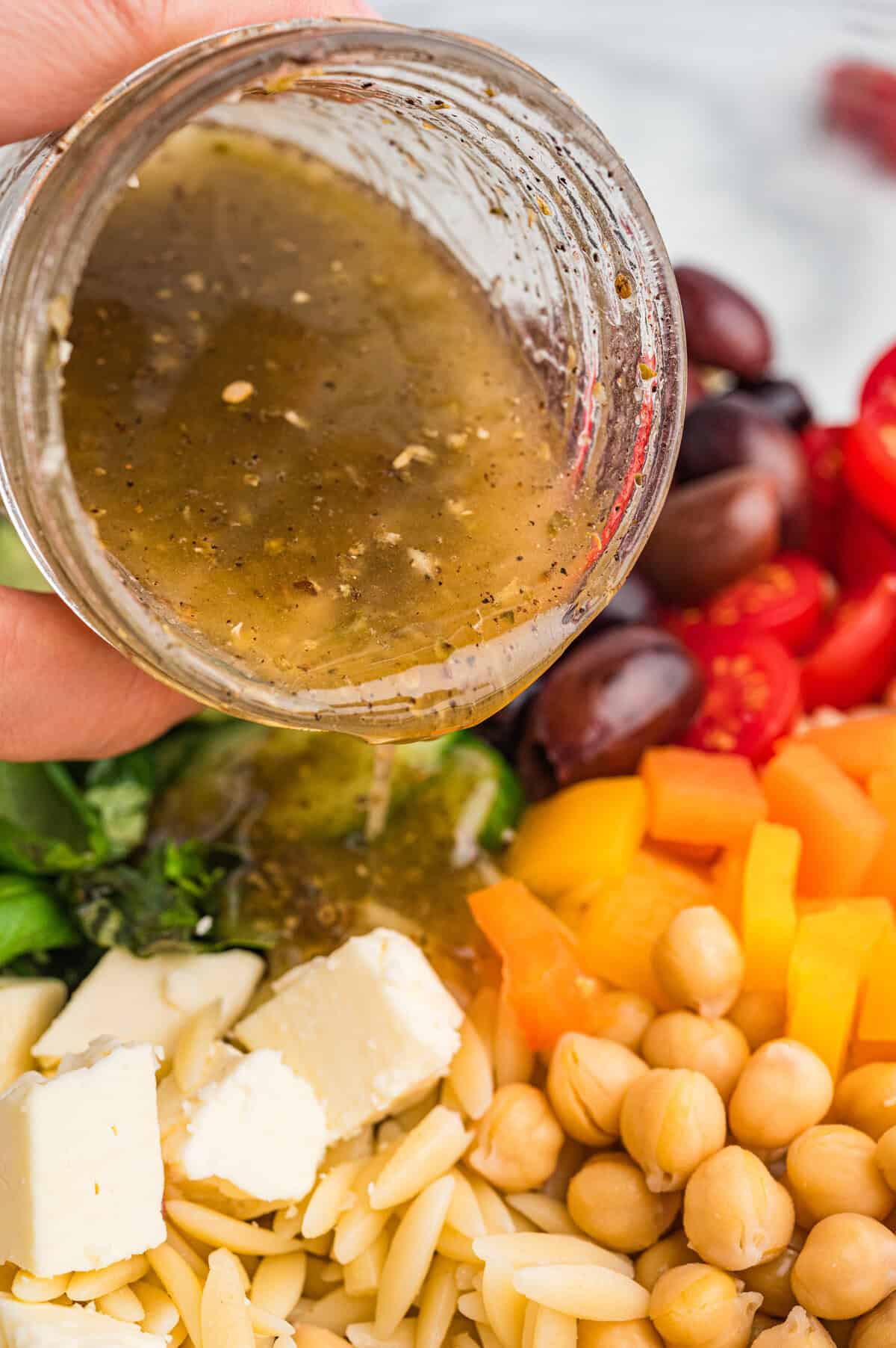 Salad dressing being poured from a jar over orzo salad ingredients.