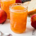 Jars of homemade peach jam with whole peaches, a spoon, and a plate with sliced bread in the background.