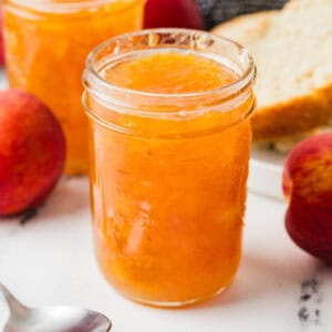 Jars of homemade peach jam with whole peaches, a spoon, and a plate with sliced bread in the background.