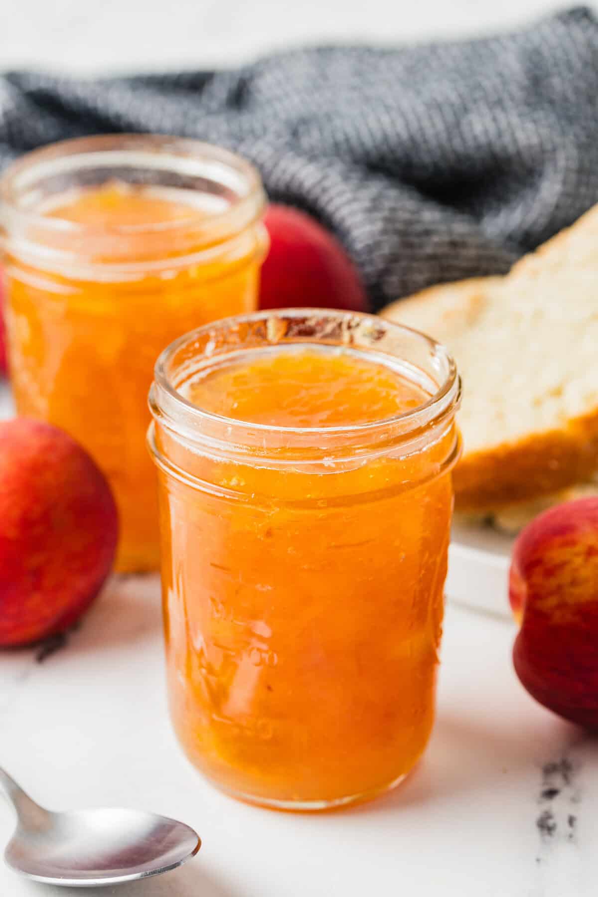 Two jars of homemade peach jam with whole peaches, a spoon, and a plate with sliced bread in the background.