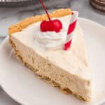 A slice of root beer float pie on a plate, topped with whipped cream, a cherry, and a red-striped paper straw.