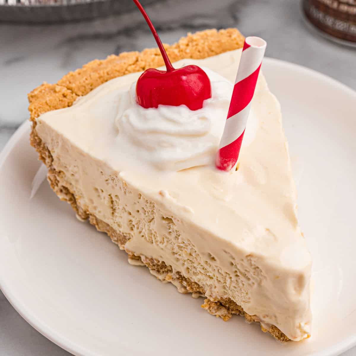 A slice of root beer float pie on a plate, topped with whipped cream, a cherry, and a red-striped paper straw.