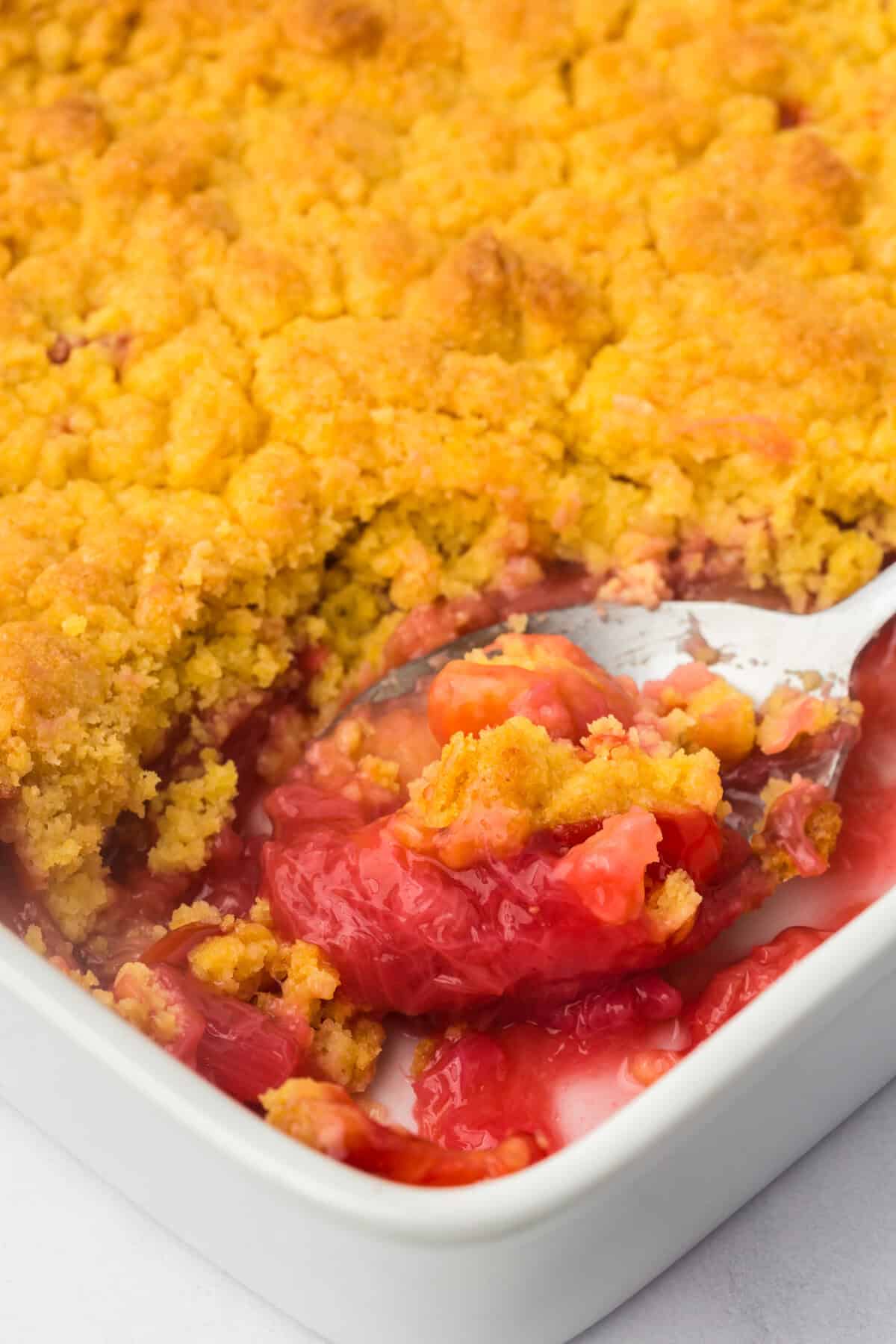 A spoon lifting a serving of strawberry rhubarb dump cake from the baking dish, showing the golden crust and bright pink-red filling.