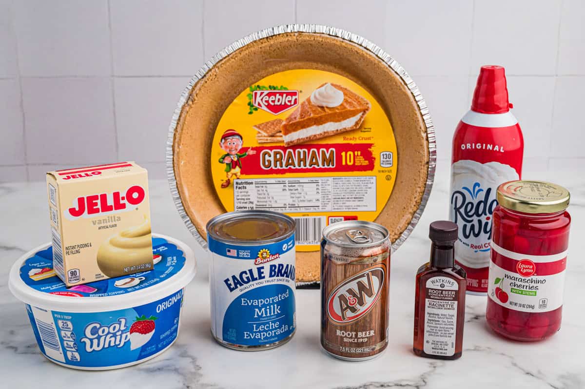 All ingredients for root beer float pie displayed: graham crust, pudding mix, Cool Whip, evaporated milk, root beer, root beer concentrae, whipped cream, and maraschino cherries.