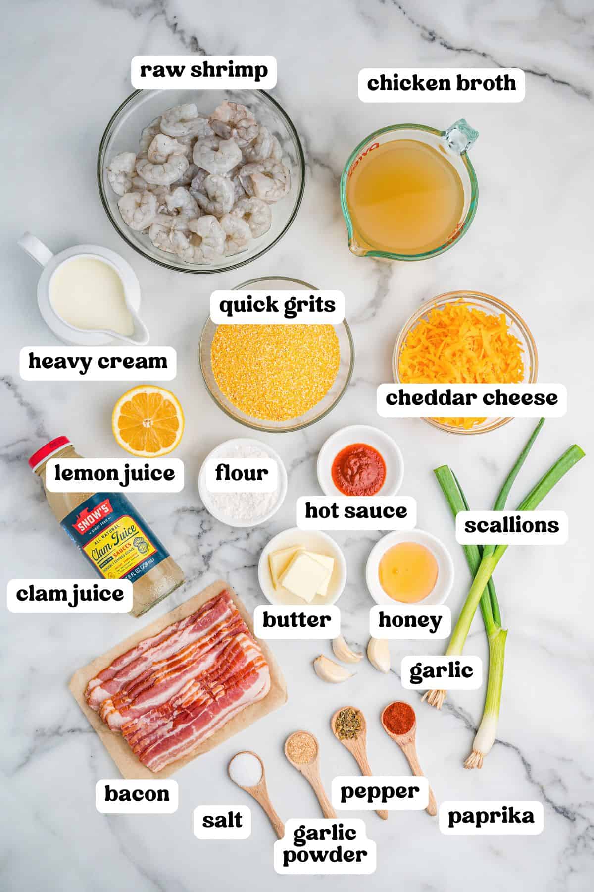 Overhead view of various labeled ingredients for cheesy shrimp and grits arranged on a marble surface. Items include raw shrimp, chicken broth, quick grits, cheddar cheese, heavy cream, lemon juice, flour, hot sauce, butter, honey, garlic, scallions, bacon, clam juice, and seasonings in wooden spoons: salt, garlic powder, pepper, and paprika.