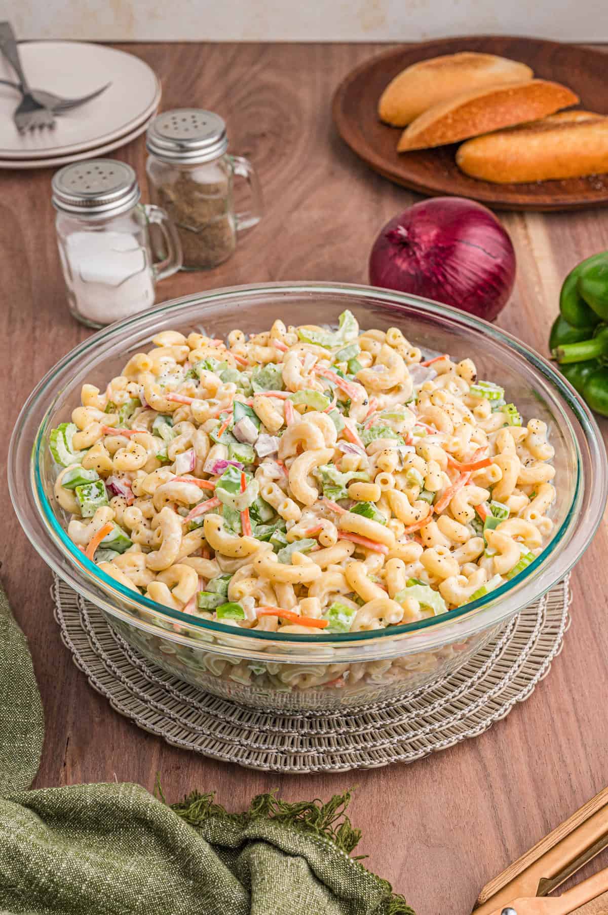 Overhead view of a large glass bowl filled with creamy macaroni salad on a wooden table. Surrounding items include salt and pepper shakers, a red onion, green bell pepper, and bread rolls.