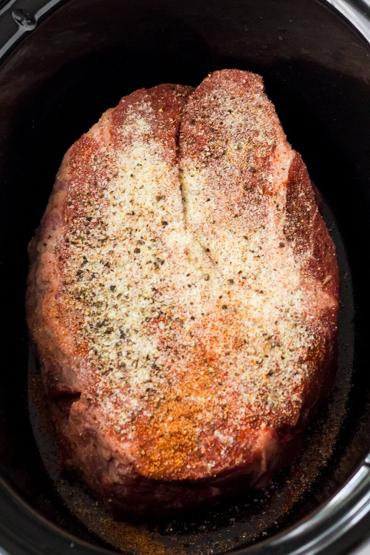 Overhead image of a large, raw chuck roast placed in a slow cooker, heavily seasoned with a dry rub of various spices including black pepper, paprika, and garlic powder.