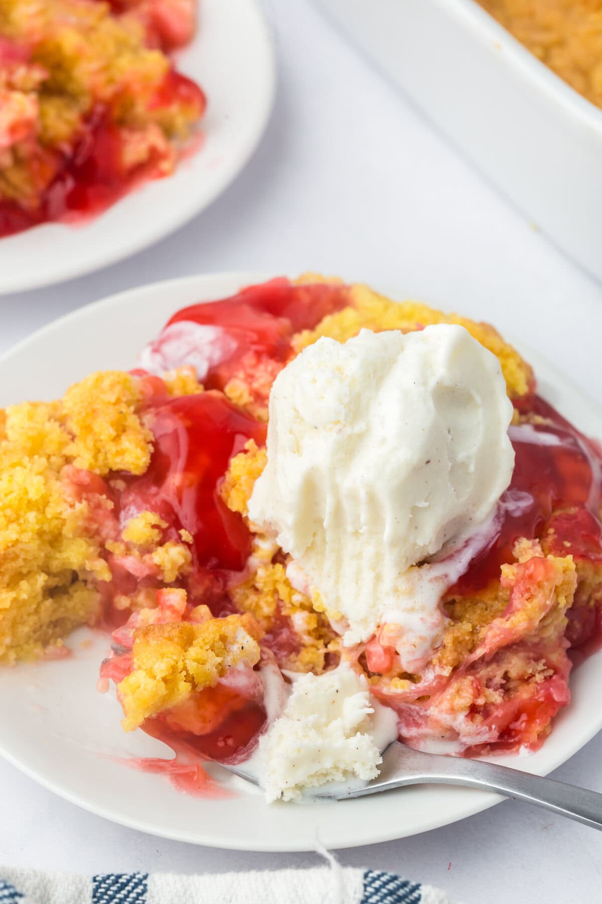 Close-up of a plate of rhubarb dump cake topped with a scoop of vanilla ice cream, showing melting cream drizzling into the cake’s red and yellow layers.