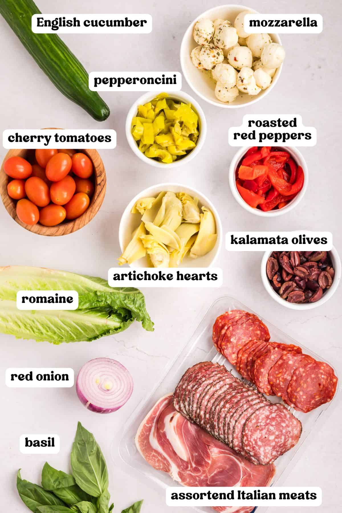 Flat lay of antipasto salad ingredients, including labeled mozzarella, pepperoncini, cherry tomatoes, roasted red peppers, kalamata olives, artichoke hearts, cucumber, red onion, romaine, basil, and assorted Italian meats.