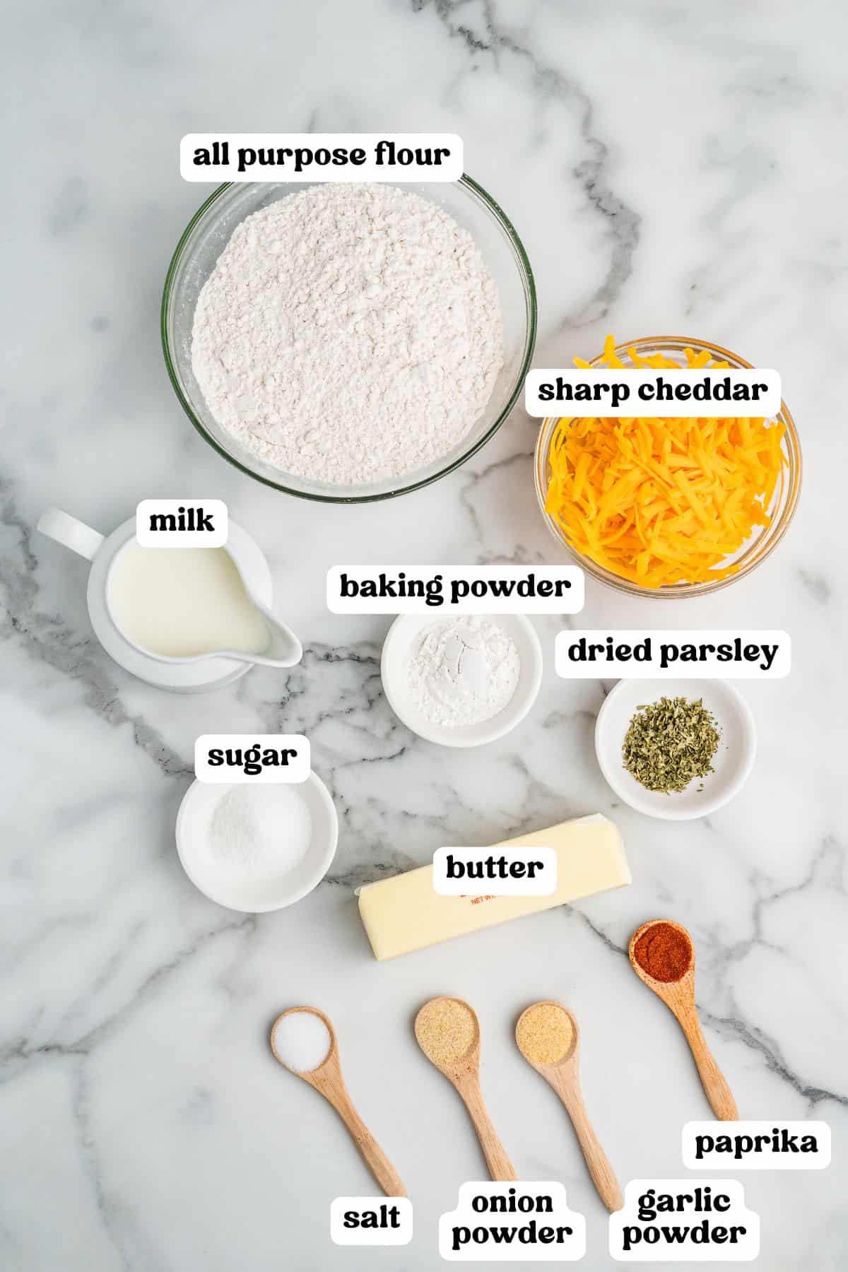 Overhead view of ingredients for cheddar biscuits arranged on a marble surface, each labeled: all-purpose flour, sharp cheddar, milk, baking powder, dried parsley, sugar, butter, salt, onion powder, garlic powder, and paprika.