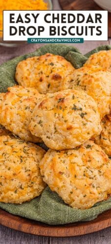A plate piled with golden cheddar drop biscuits on a green cloth. Text at the top reads “Easy Cheddar Drop Biscuits” with the site name “crayonsandcravings.com.”