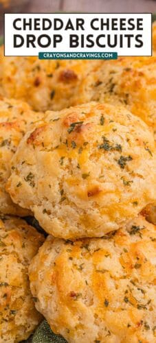 Close-up of several golden cheddar drop biscuits topped with herbs. Bold text at the top reads “Cheddar Cheese Drop Biscuits” with the website “crayonsandcravings.com.”